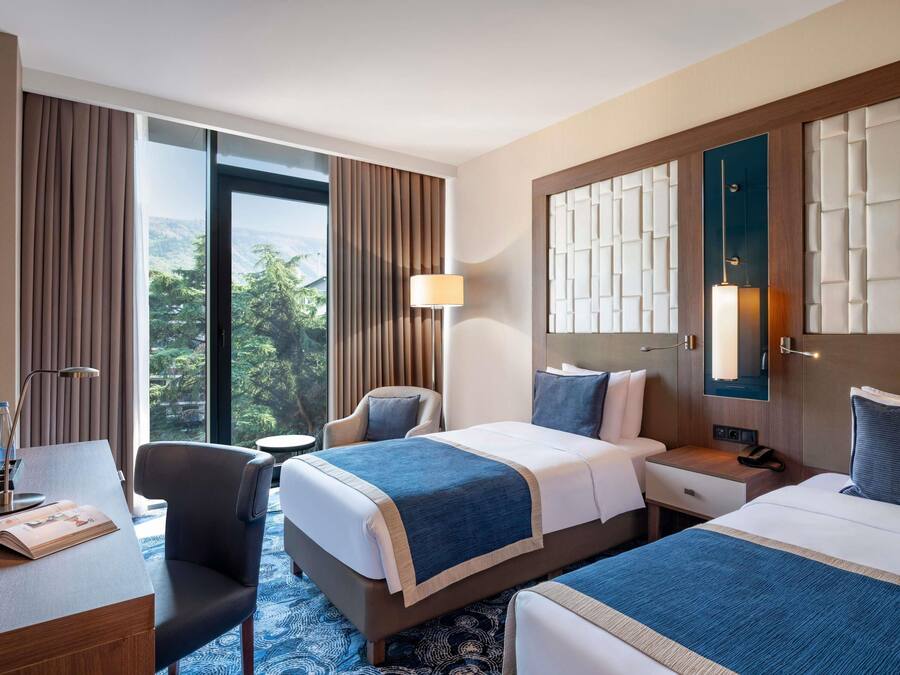 A hotel room with two beds, a desk, a chair, a lamp, and a view of greenery.
