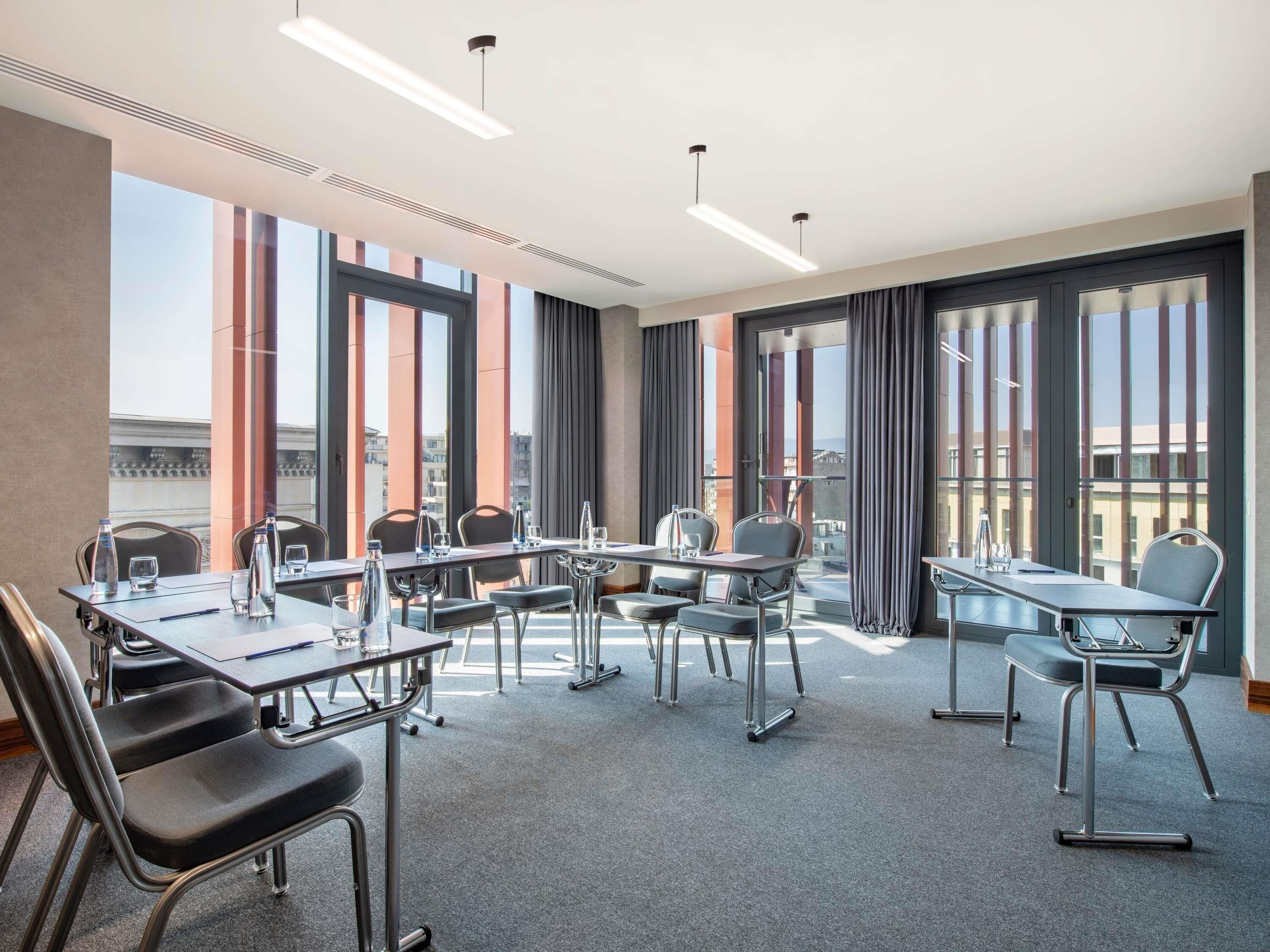 A conference room with tables and chairs, large windows with curtains, and a view of buildings outside.