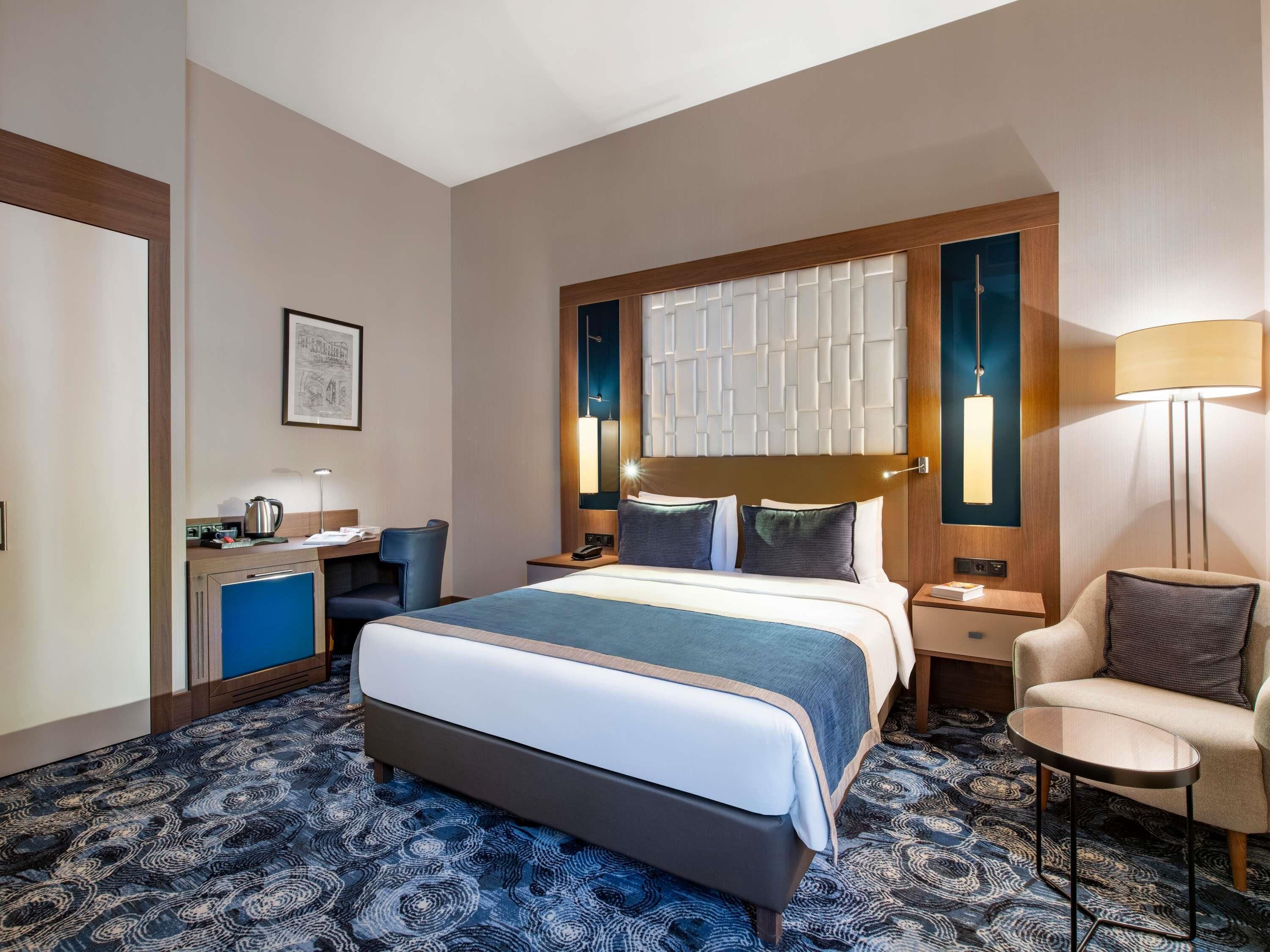 A hotel room with a bed, desk, chair, and a lamp.