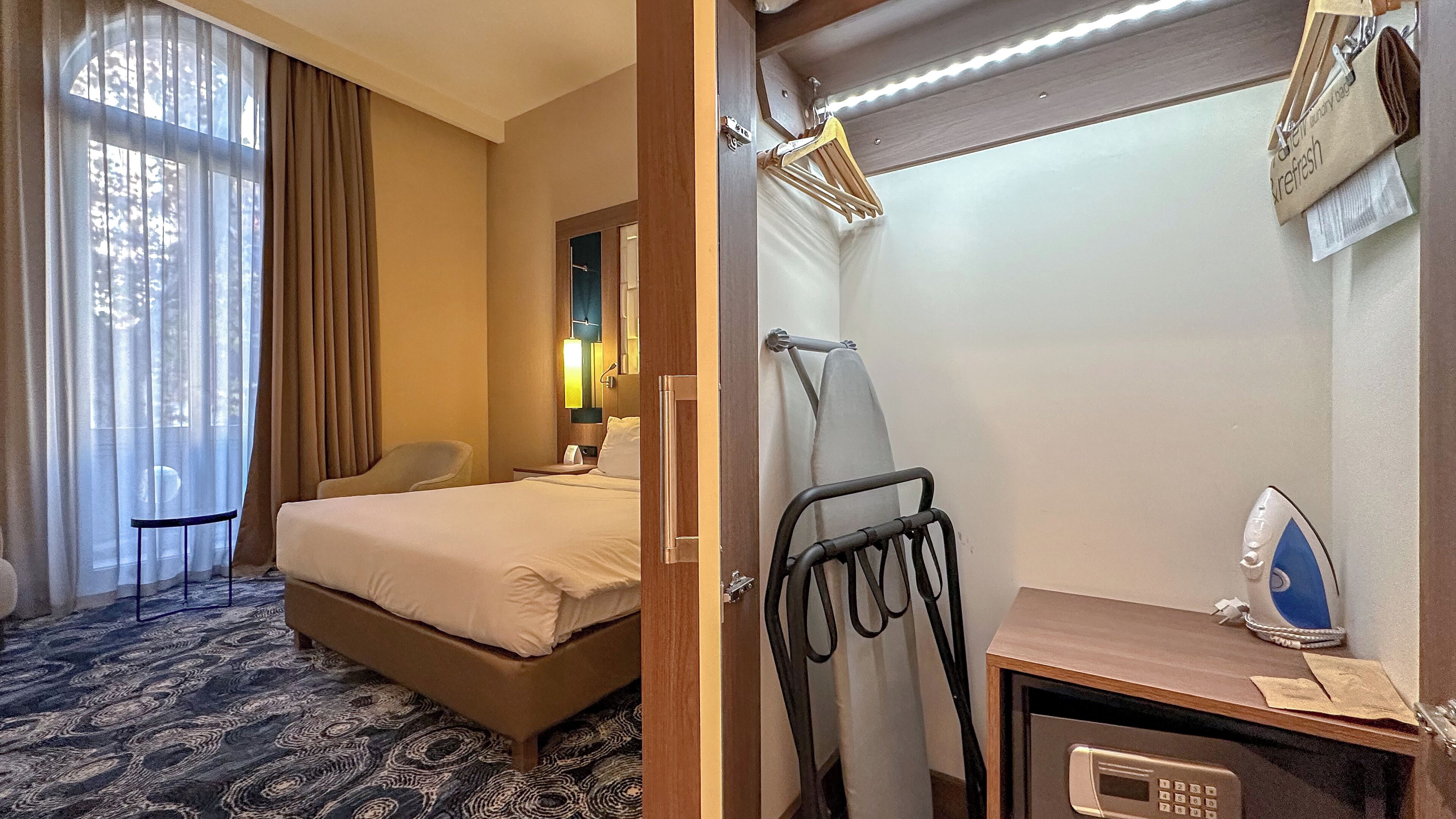 A hotel room with a bed, a chair, a desk with a microwave, and a hanging clothes rack.