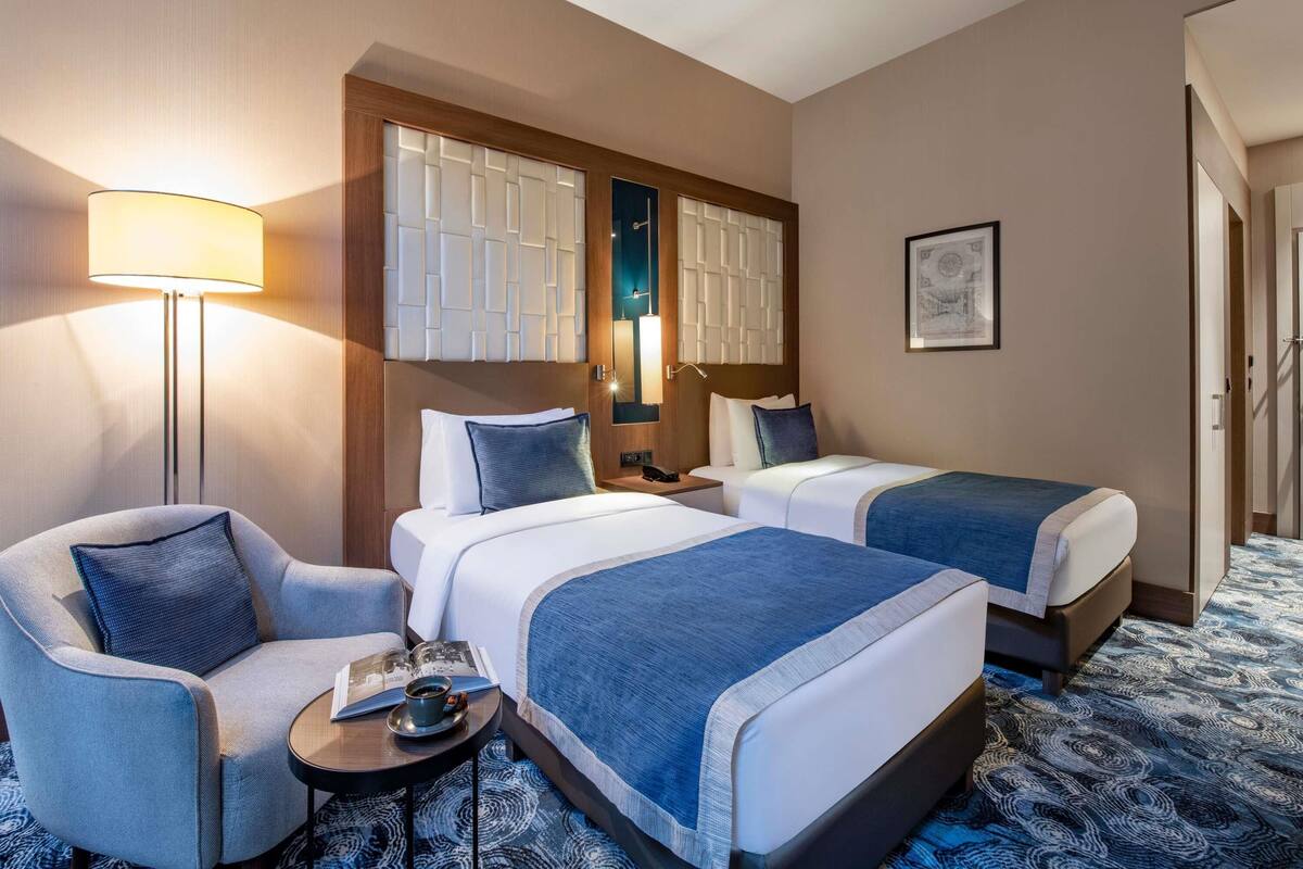 A hotel room with two beds, a chair, a small table with a cup and book, and a lamp.