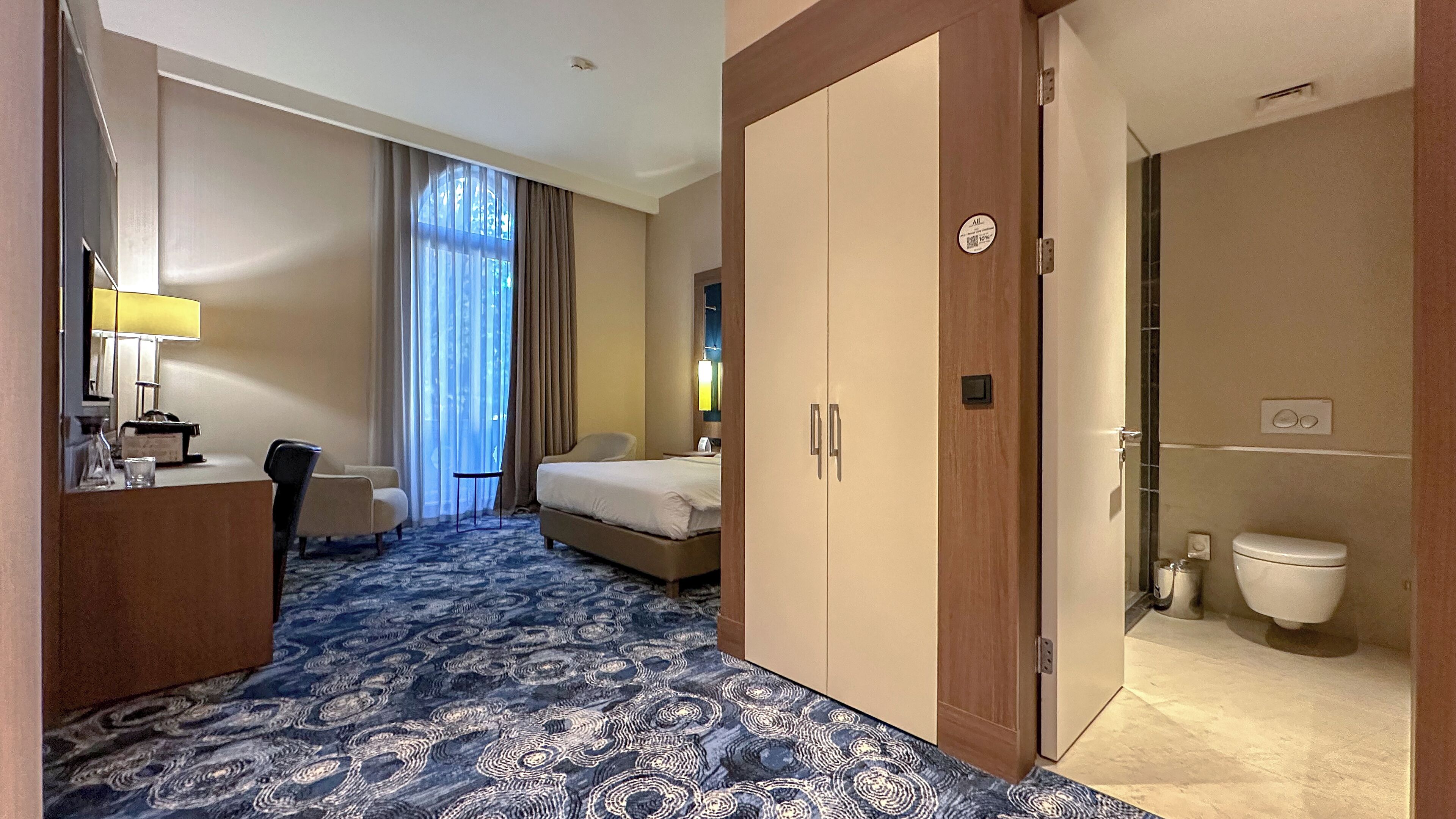 Hotelkamer met een bed, een bureau met lamp, een stoel, een badkamer met toilet en een groot raam met gordijnen.