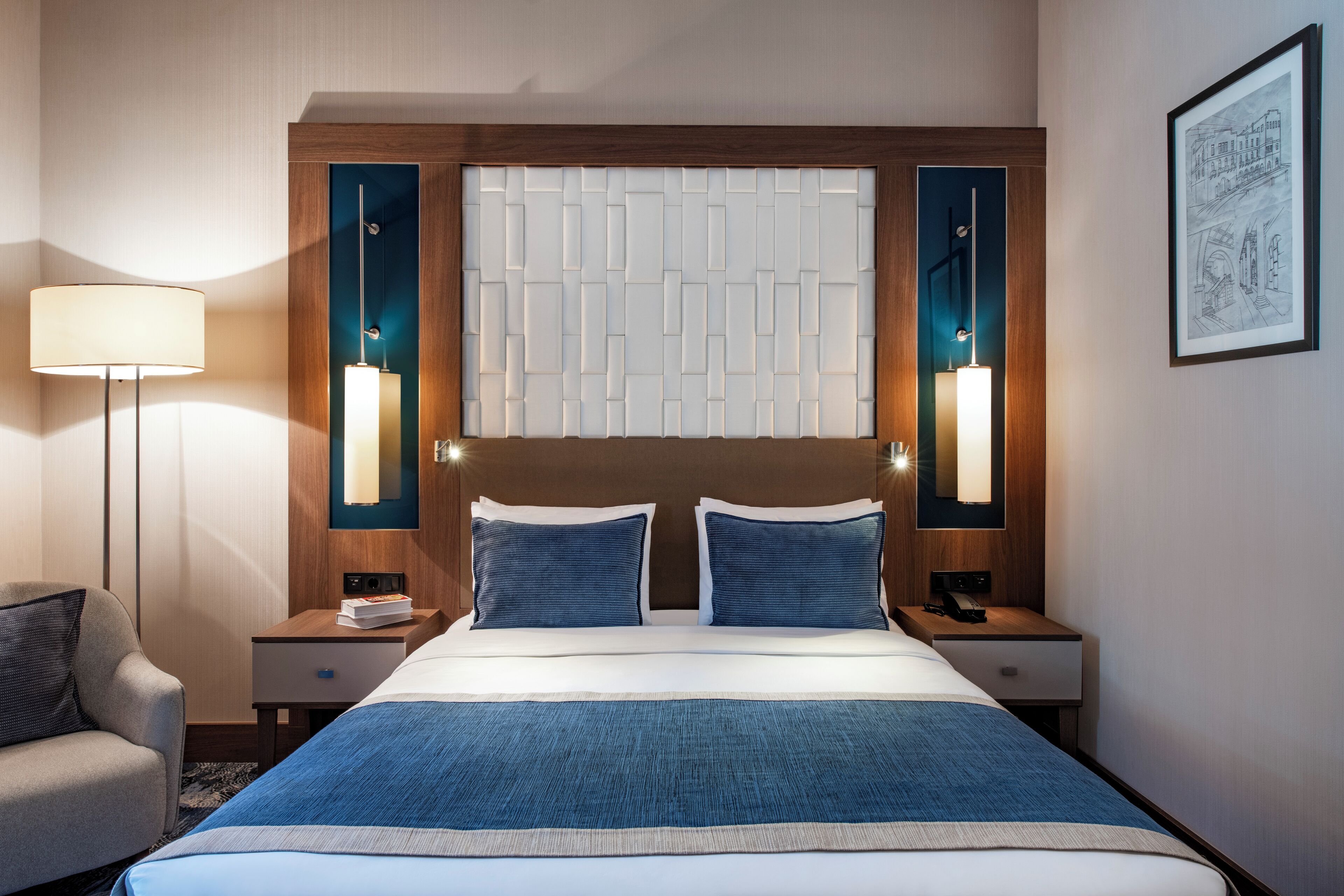 A hotel room with a large bed, two bedside tables, a chair, a wall-mounted lamp, and a framed picture.