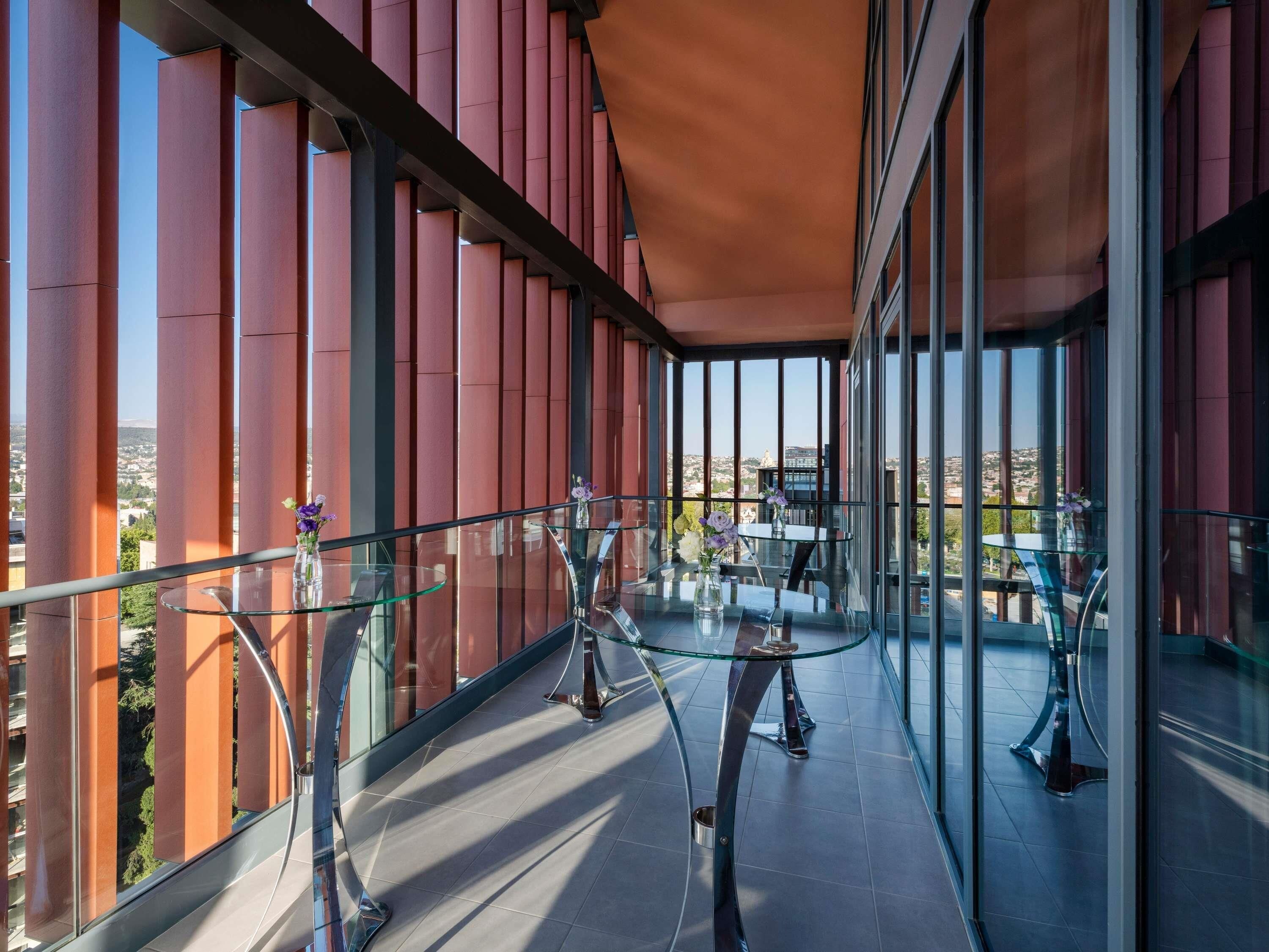 A modern balcony with glass railings, a glass table, and chairs, overlooking a cityscape.