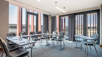 Un salón de conferencias con mesas y sillas, amplias ventanas con cortinas y vistas a edificios.