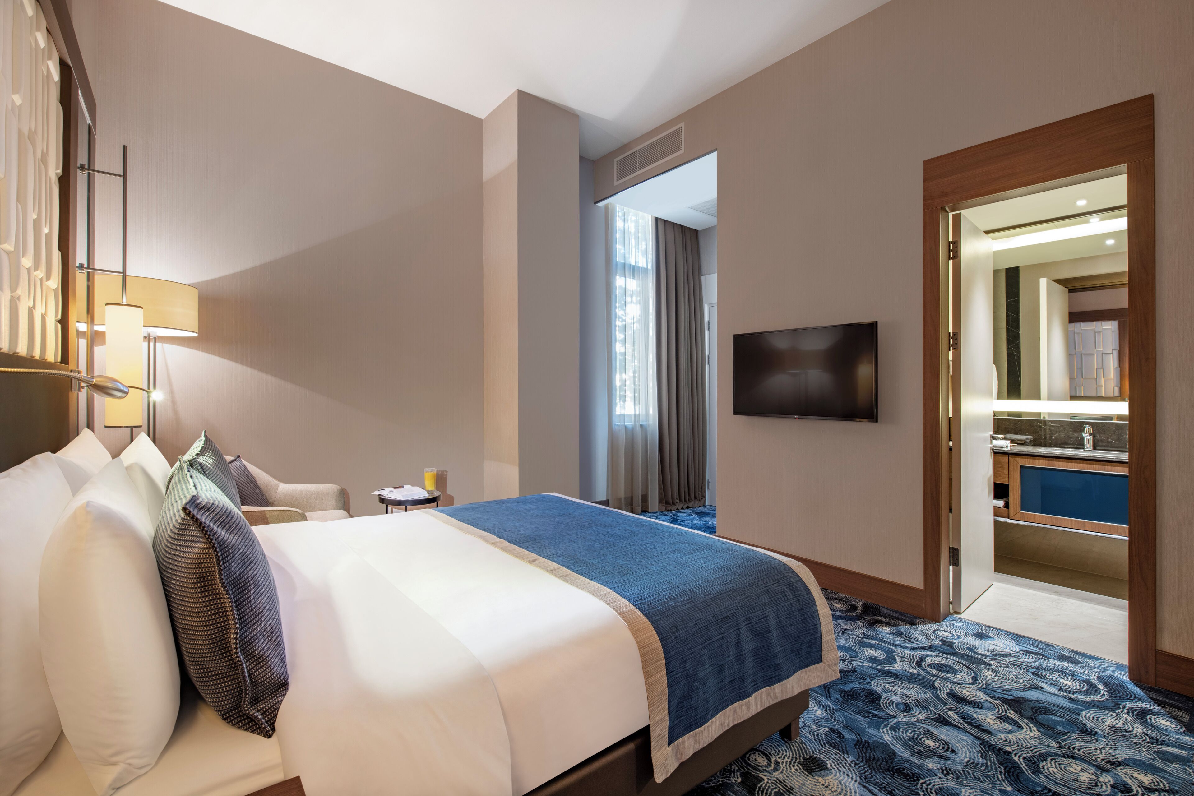 A hotel room with a bed, a TV, a desk, and a bathroom visible through an open door.