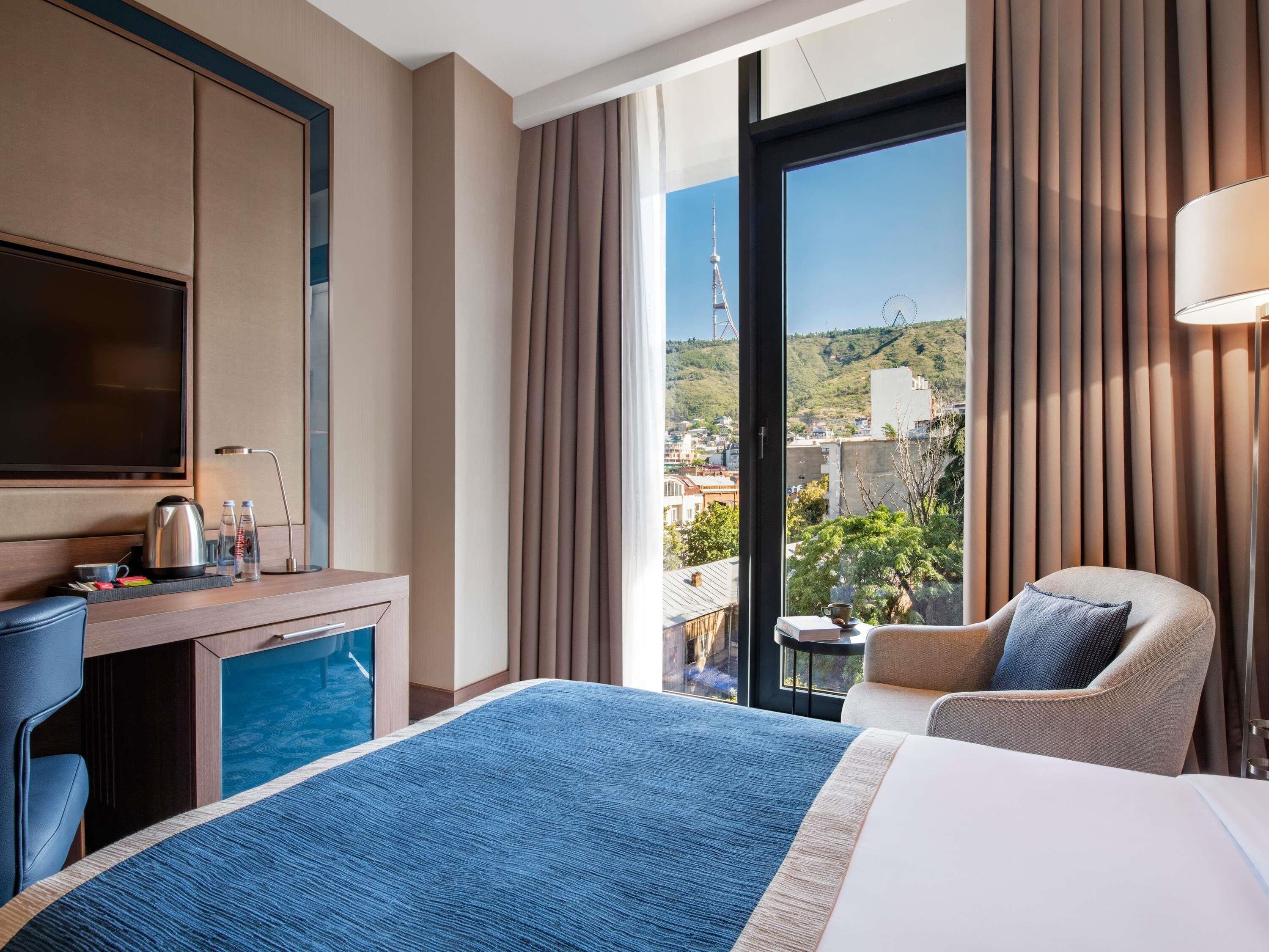 A hotel room with a bed, a desk with a kettle, a chair, a TV, and a view of a cityscape.
