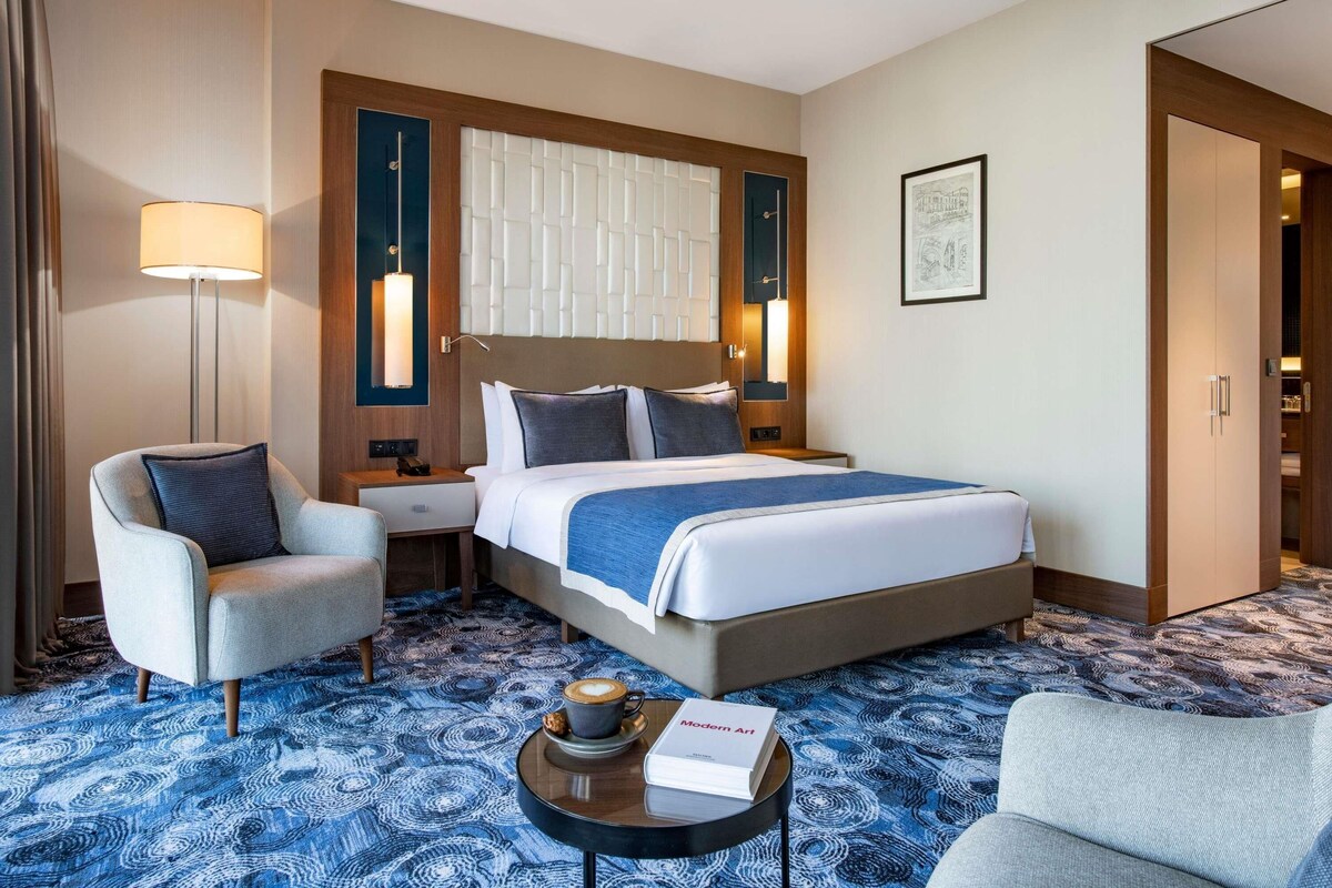 A hotel room with a bed, two bedside tables, a chair, a coffee table, and a lamp.