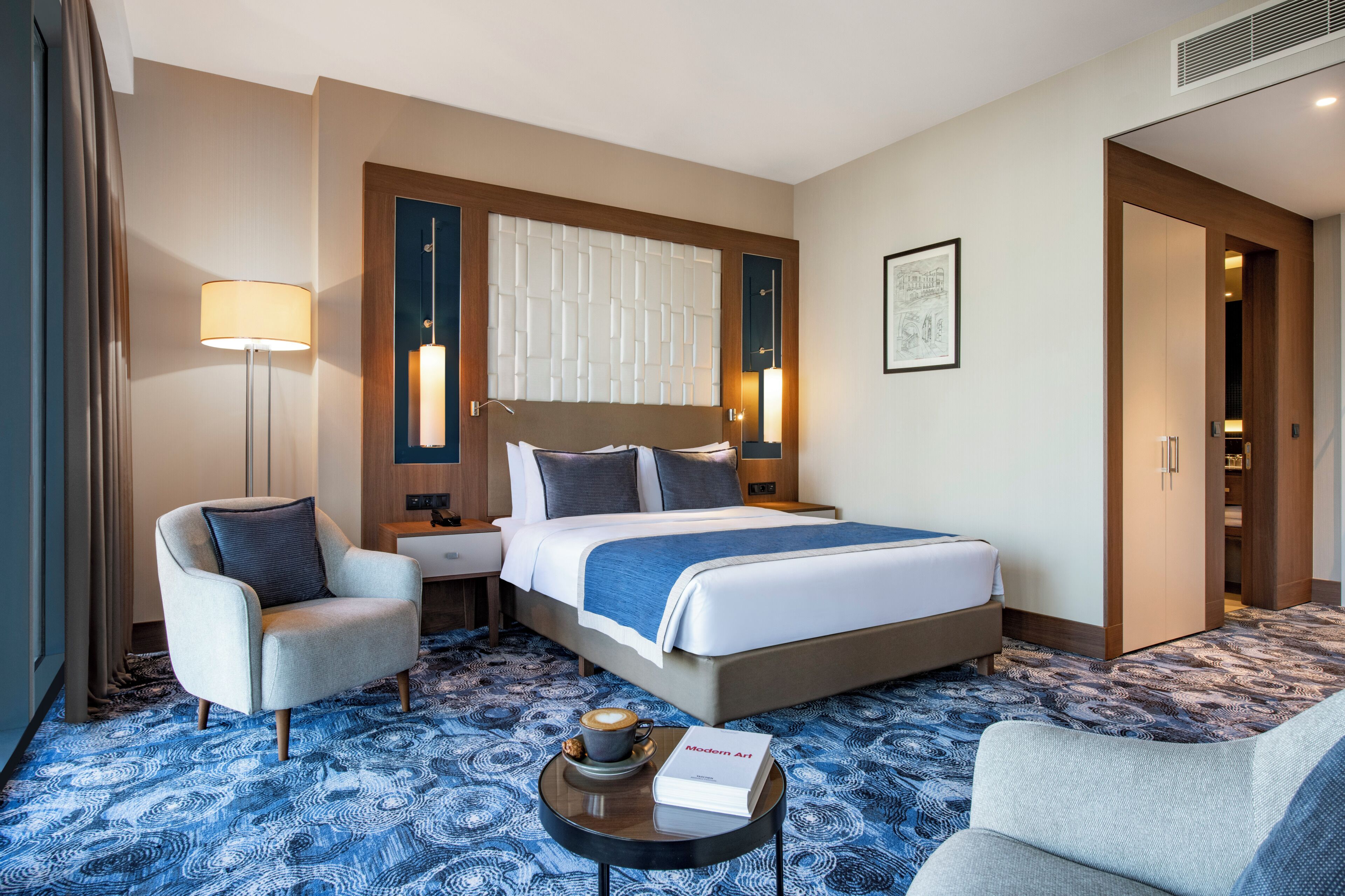 A hotel room with a large bed, two armchairs, a coffee table, and a blue patterned carpet.