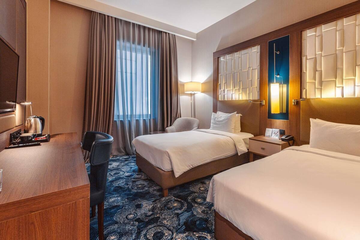 A hotel room with a bed, bedside tables, a desk, a chair, a lamp, and a window with curtains.
