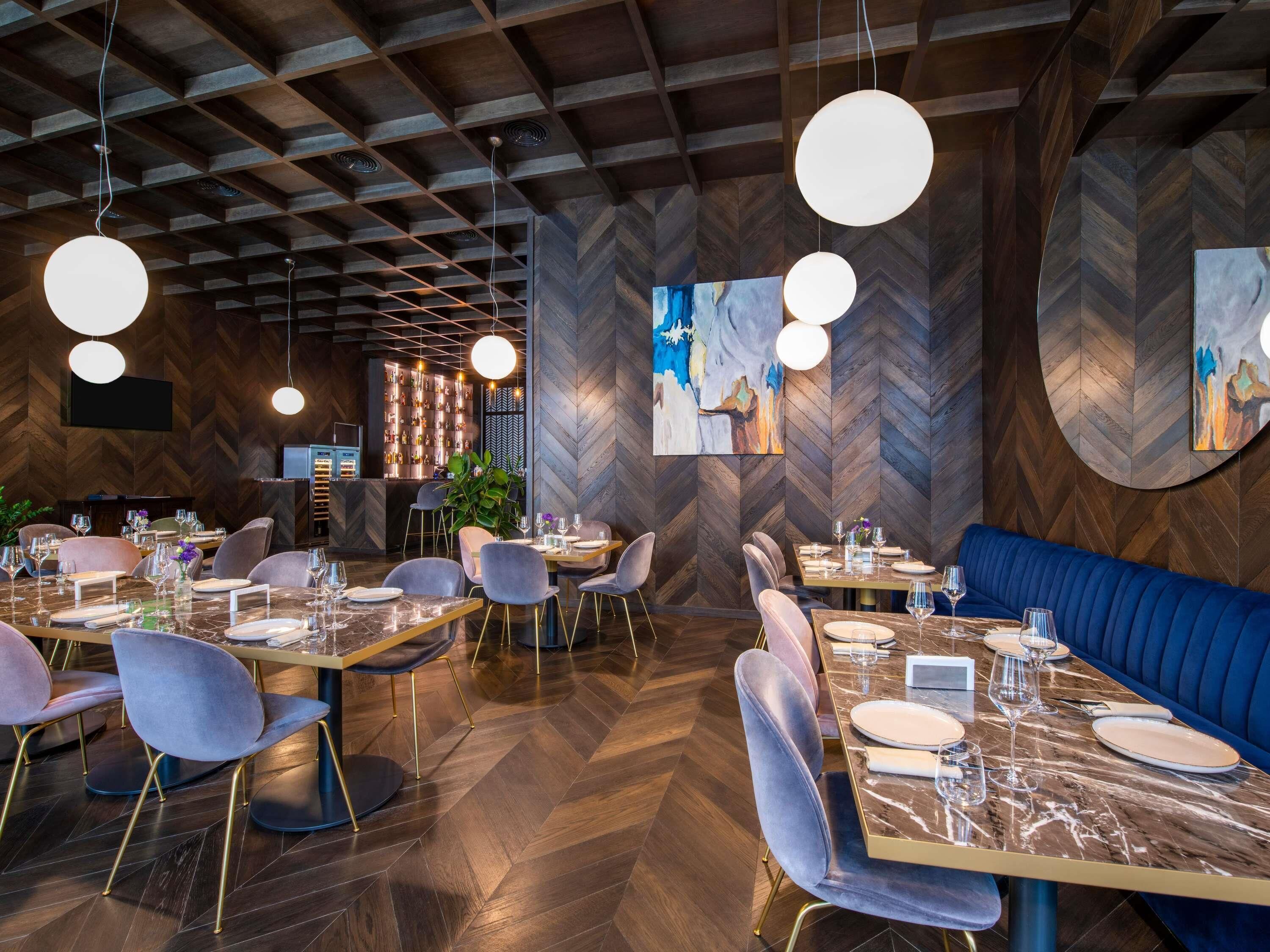 A restaurant interior with herringbone wood flooring, round pendant lighting, and a blue upholstered booth.