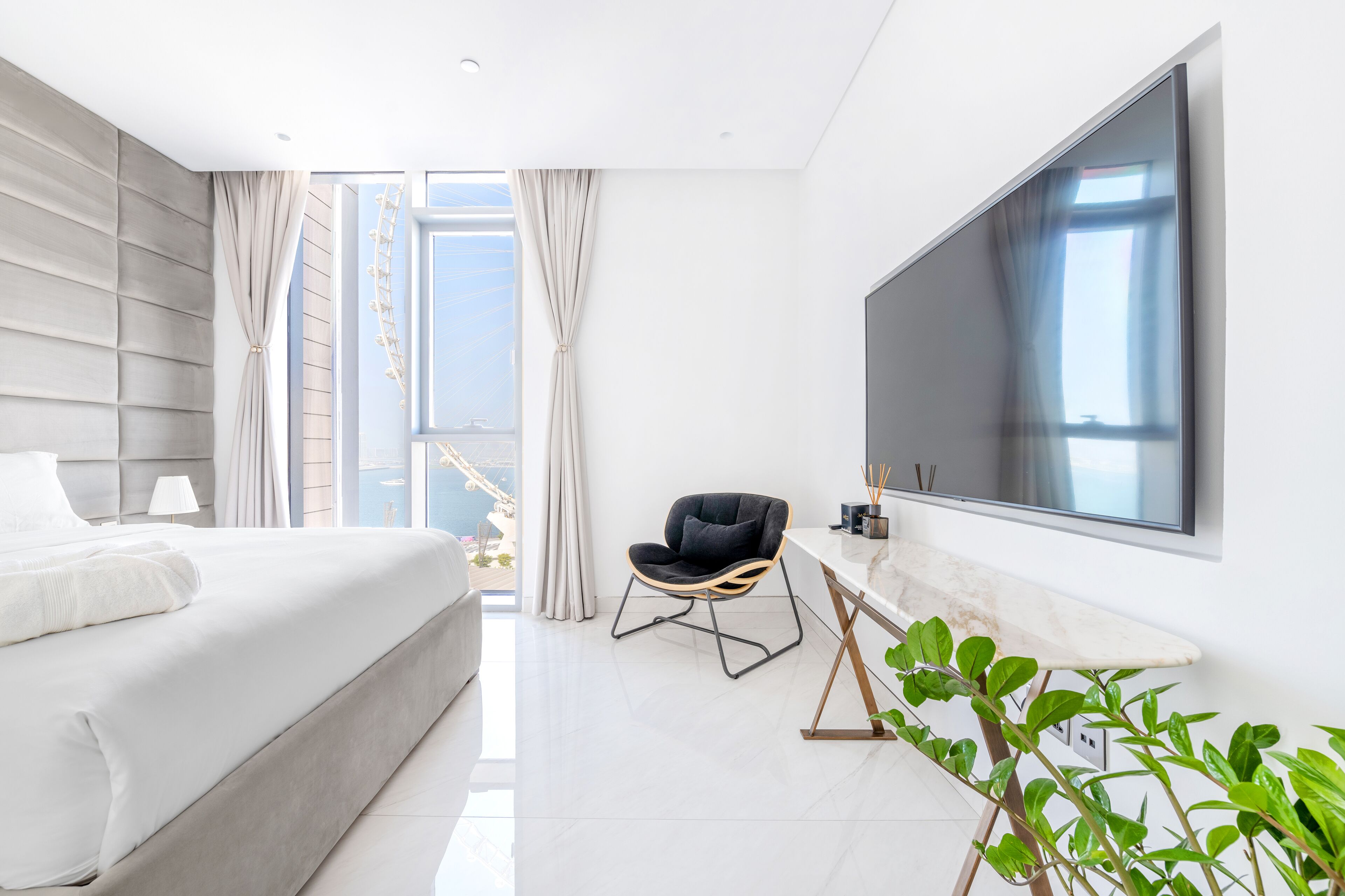 A modern hotel room with a large bed, a seating area with a chair, a flat-screen TV, and a plant.
