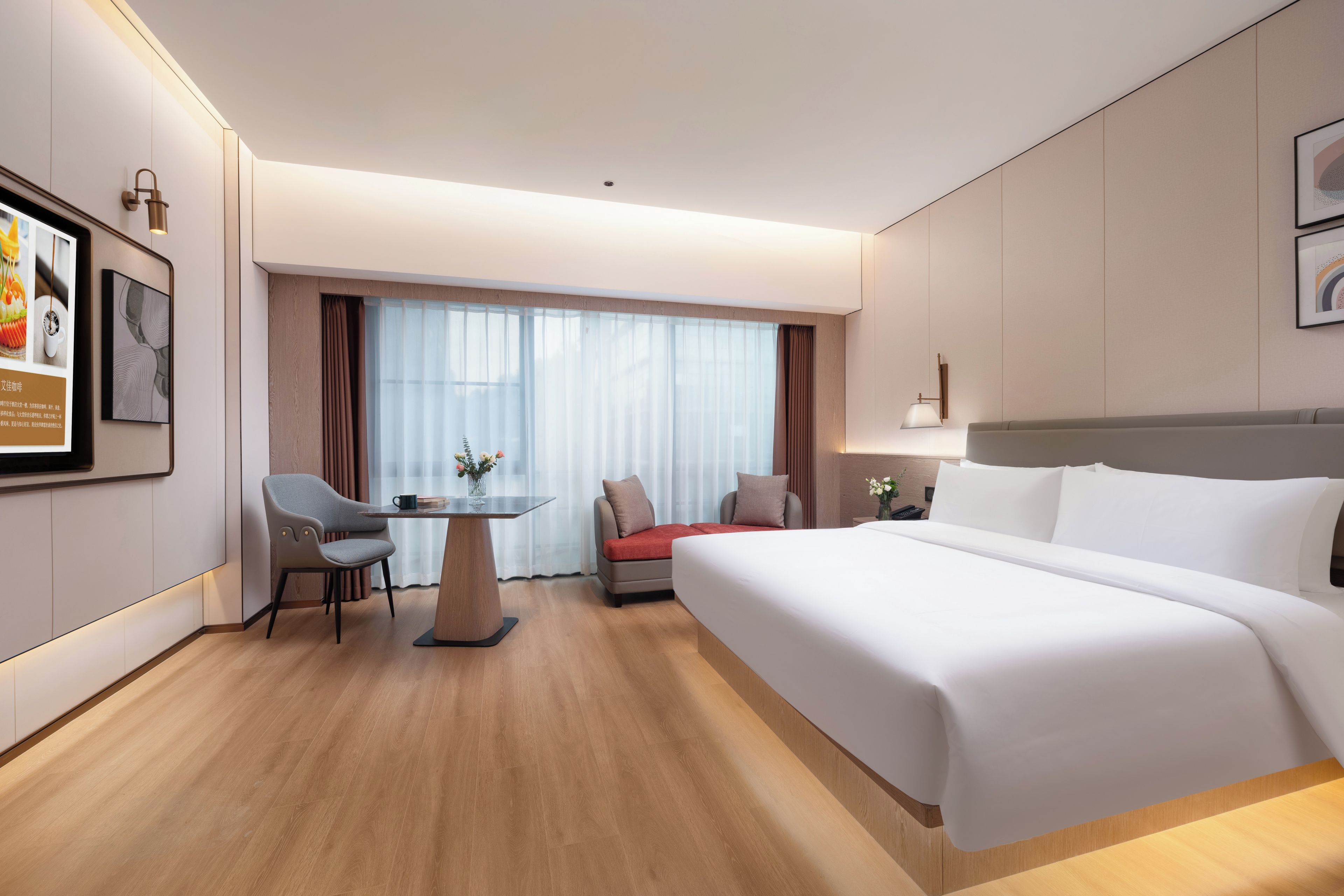 Ein Hotelzimmer mit einem großen Bett, einem Schreibtisch mit Stuhl, einem kleinen Tisch mit Blumenvase und einem Fenster mit Vorhängen.