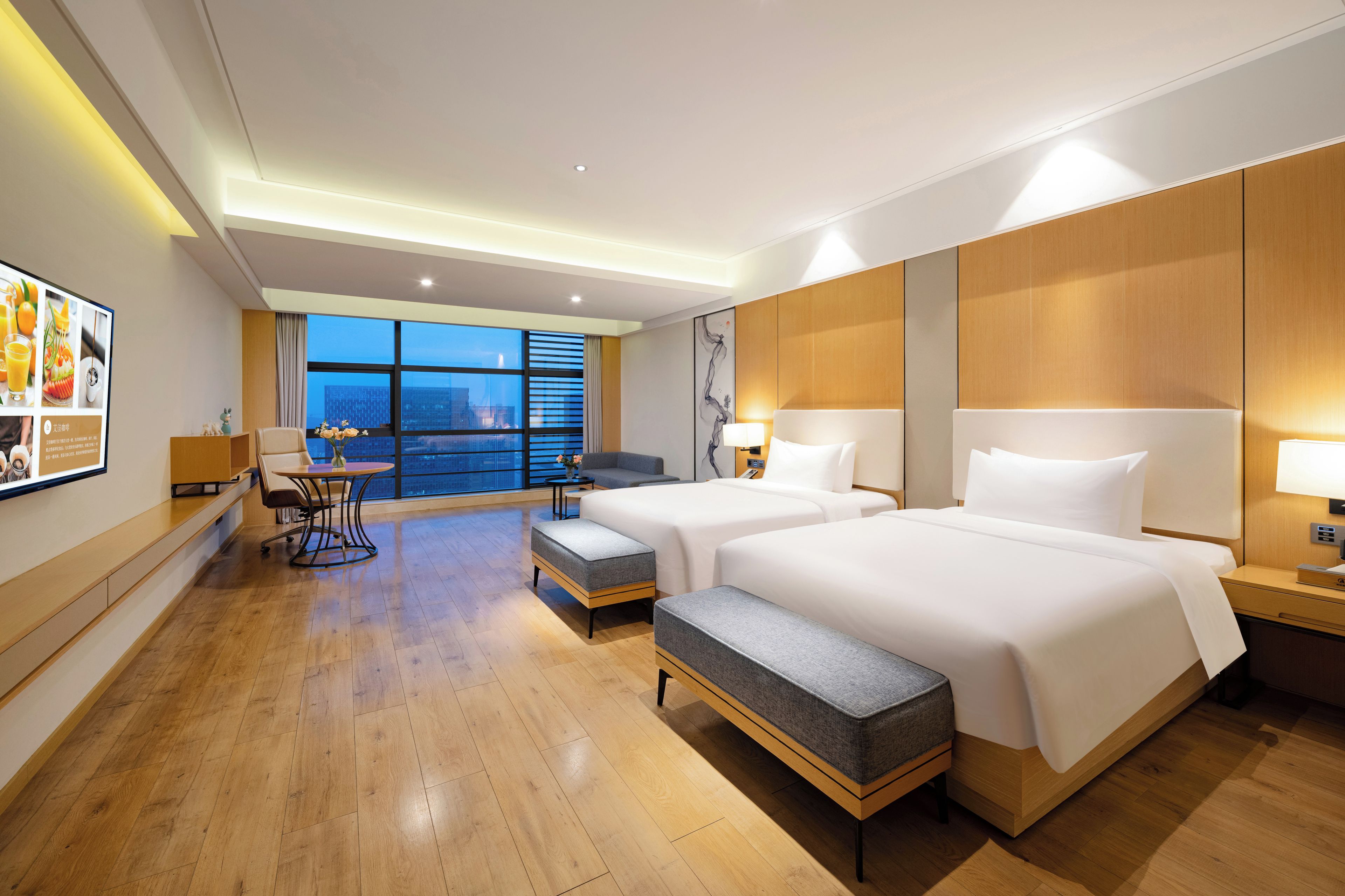 A hotel room with two beds, a desk, a chair, a small table, and a large window offering a city view.