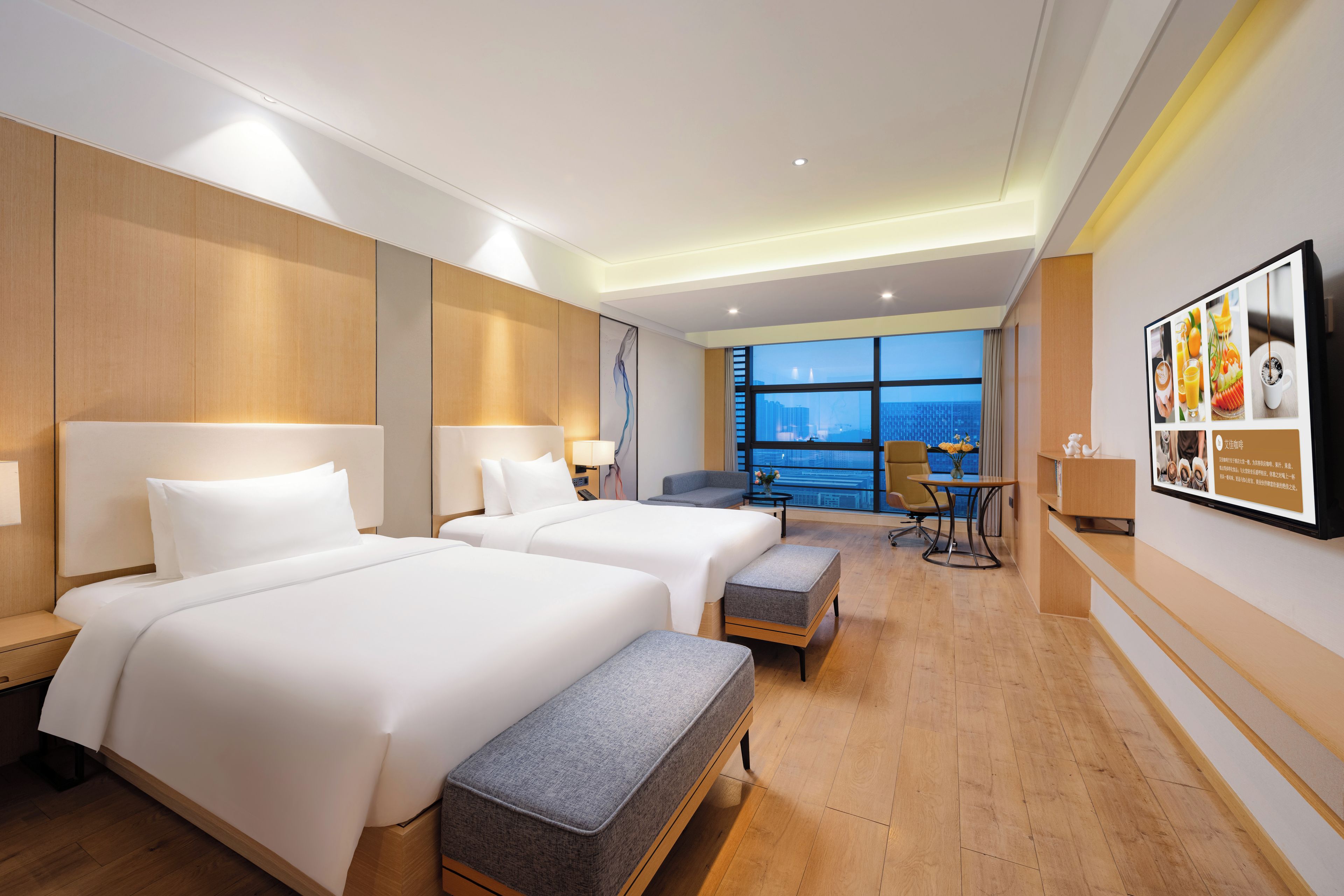 Habitación de hotel con dos camas, zona de estar, mesa de comedor y un amplio ventanal con vistas a la ciudad.