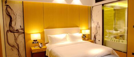 A modern hotel room with a large bed, bedside lamps, a nightstand, and a glass-enclosed bathroom.