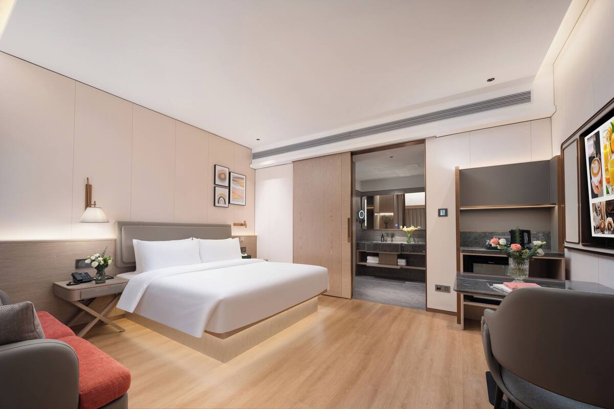 Ett modernt hotellrum med en stor säng, ett skrivbord, en stol, ett nattduksbord och ett badrum som syns genom en öppen dörr.