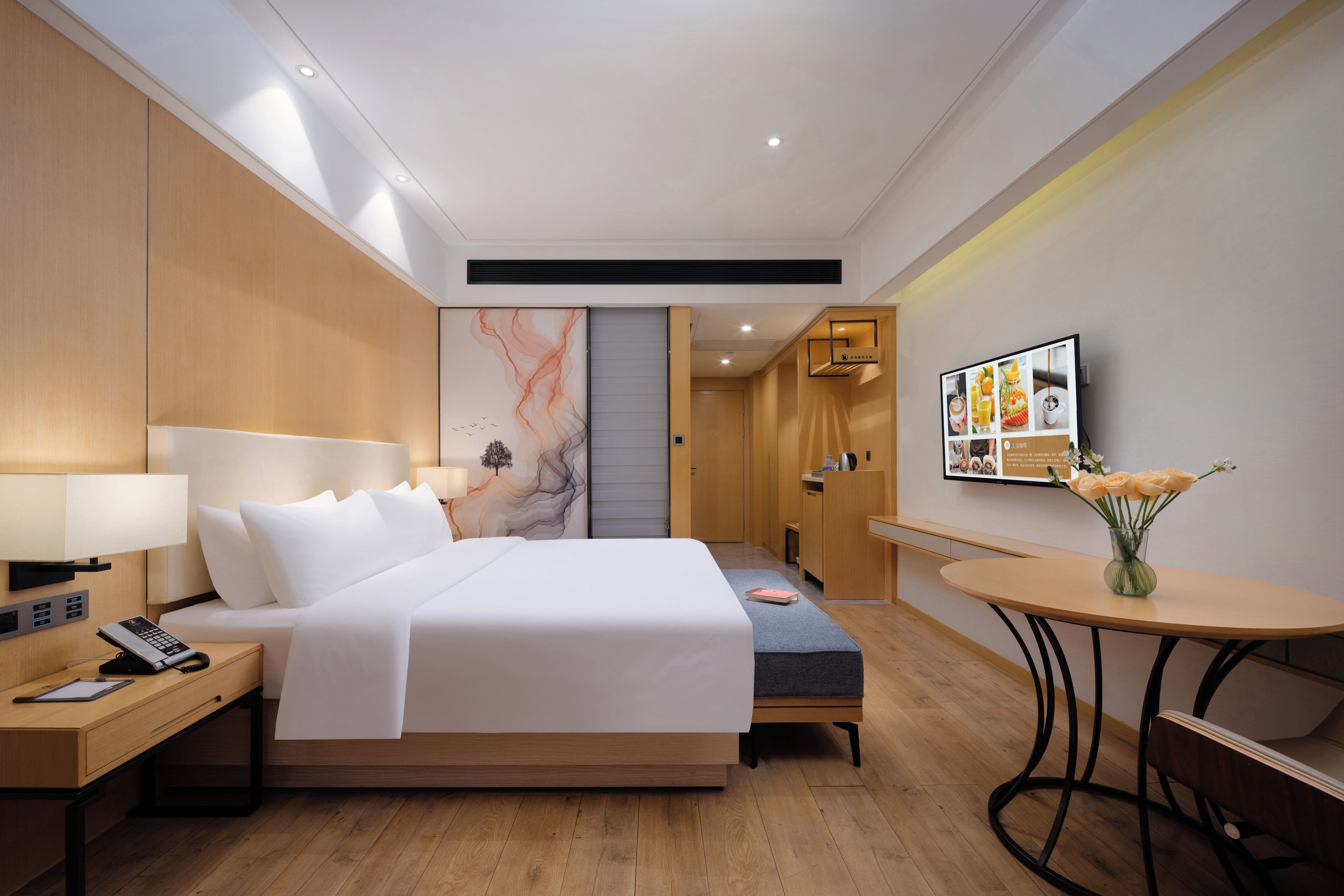 A hotel room with a bed, bedside table, lamp, desk, chair, and a small round table with a vase of flowers.