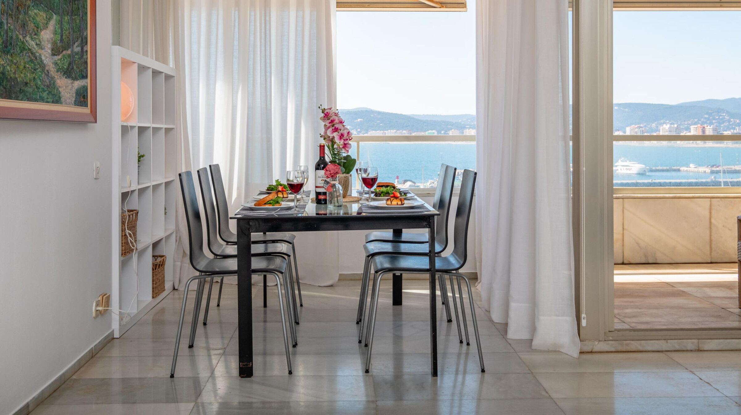 Un comedor con una mesa dispuesta para dos personas, sillas, una botella de vino y vistas al mar.