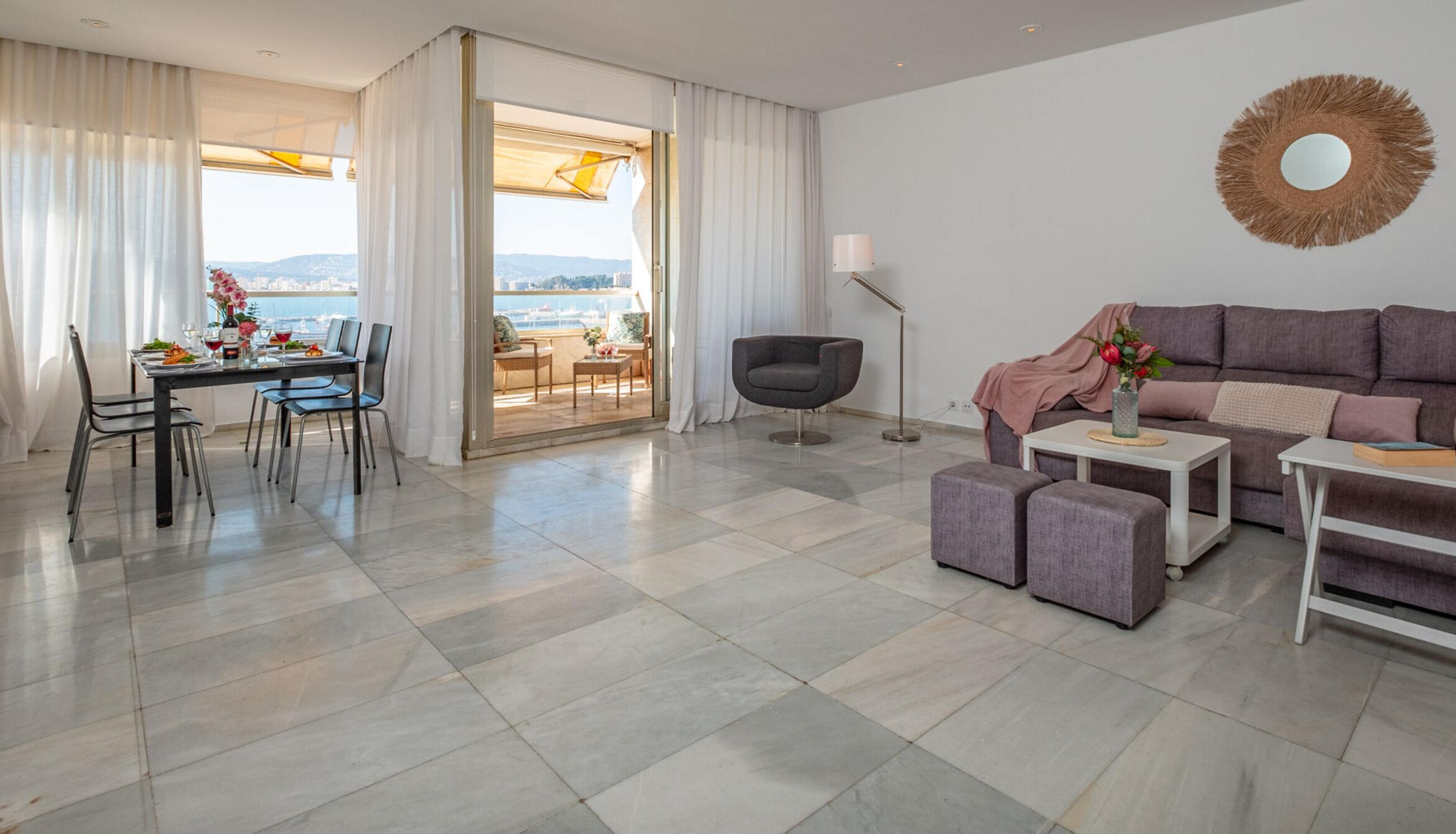 Una sala moderna con un sofá gris, un comedor con mesa dispuesta para dos personas, y un balcón con vista a la ciudad.