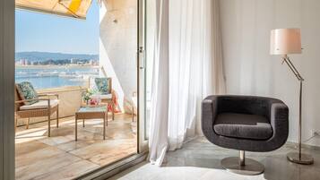 Habitación moderna con vistas a un paisaje urbano y al agua. Cuenta con una zona de estar con mobiliario de mimbre, un sillón oscuro y una lámpara de pie.