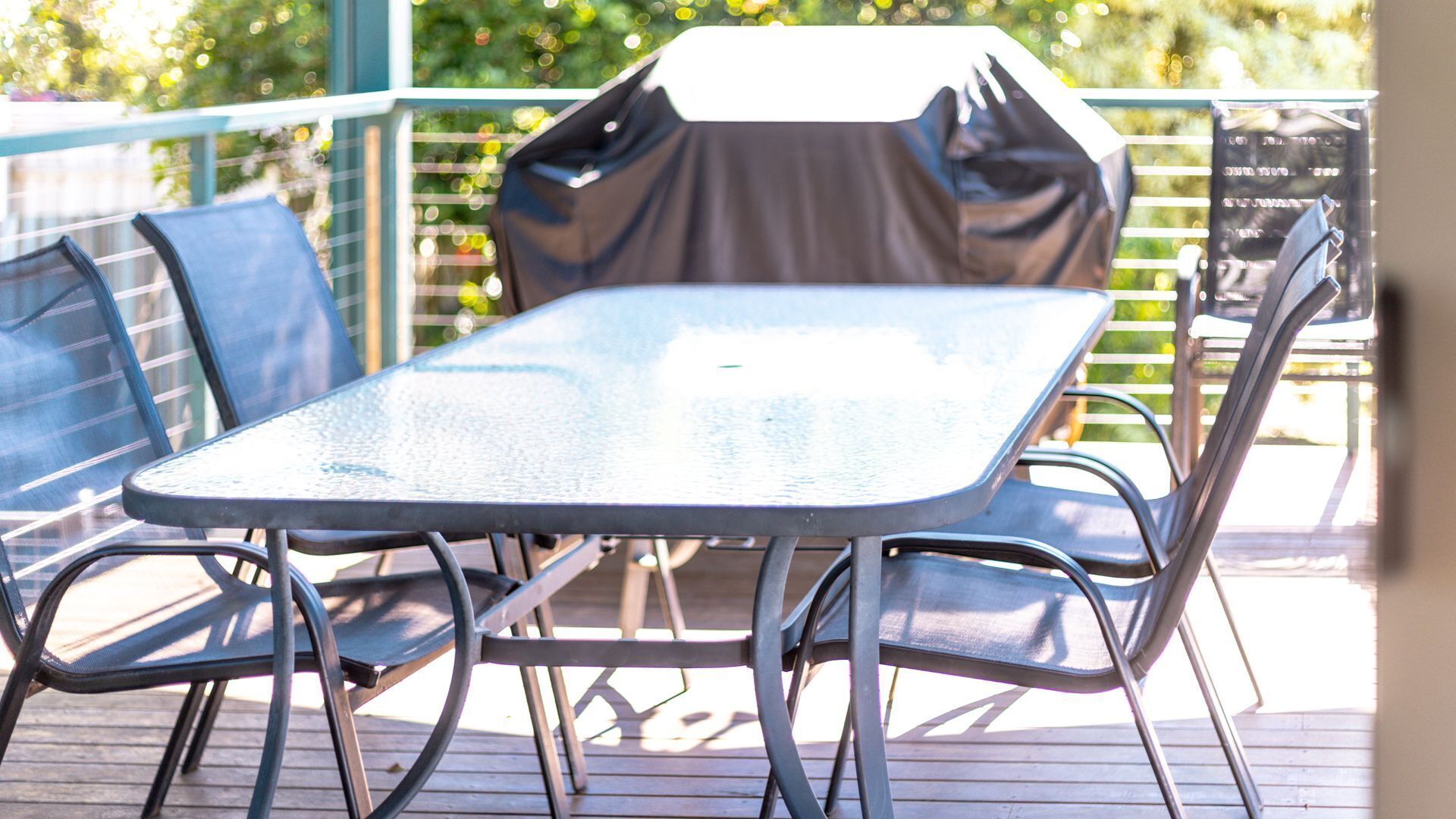 A metal table and chairs on an outdoor deck with a grill in the background.