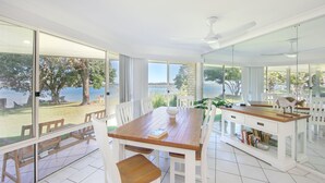 A modern dining area with a wooden table and white chairs, large windows offering a view of a garden and a lake, and a glass partition separating the dining area from a kitchen area. - River Luxury - Set right on Iluka Bay (Iluka)