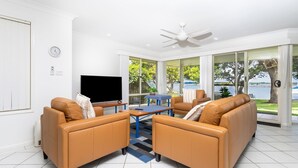 A modern living room with leather sofas, a flat-screen TV, and large windows offering a view of a body of water. - River Luxury - Set right on Iluka Bay (Iluka)