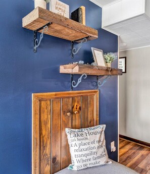 A room with a dark blue wall, wooden headboard, and a pillow with text. There are two wooden shelves with metal brackets, one holding books and a sign that says "Hook Wine On".