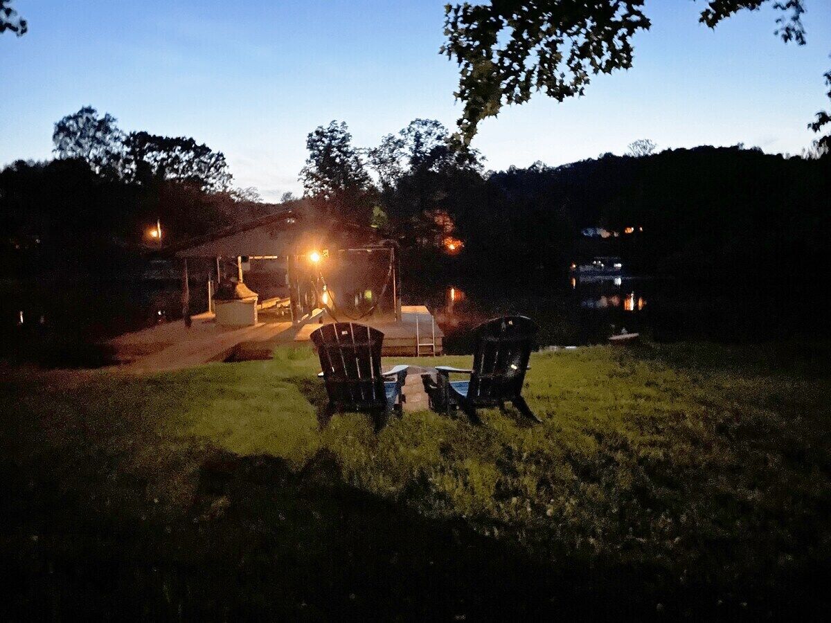 A grassy outdoor area with two chairs and a table, a vehicle with lights on, and a building in the background.