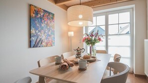 Ein Essbereich mit einem Holztisch, weißen Stühlen, einem Bild an der Wand, einer Vase mit Blumen und einem Fenster mit Jalousien.