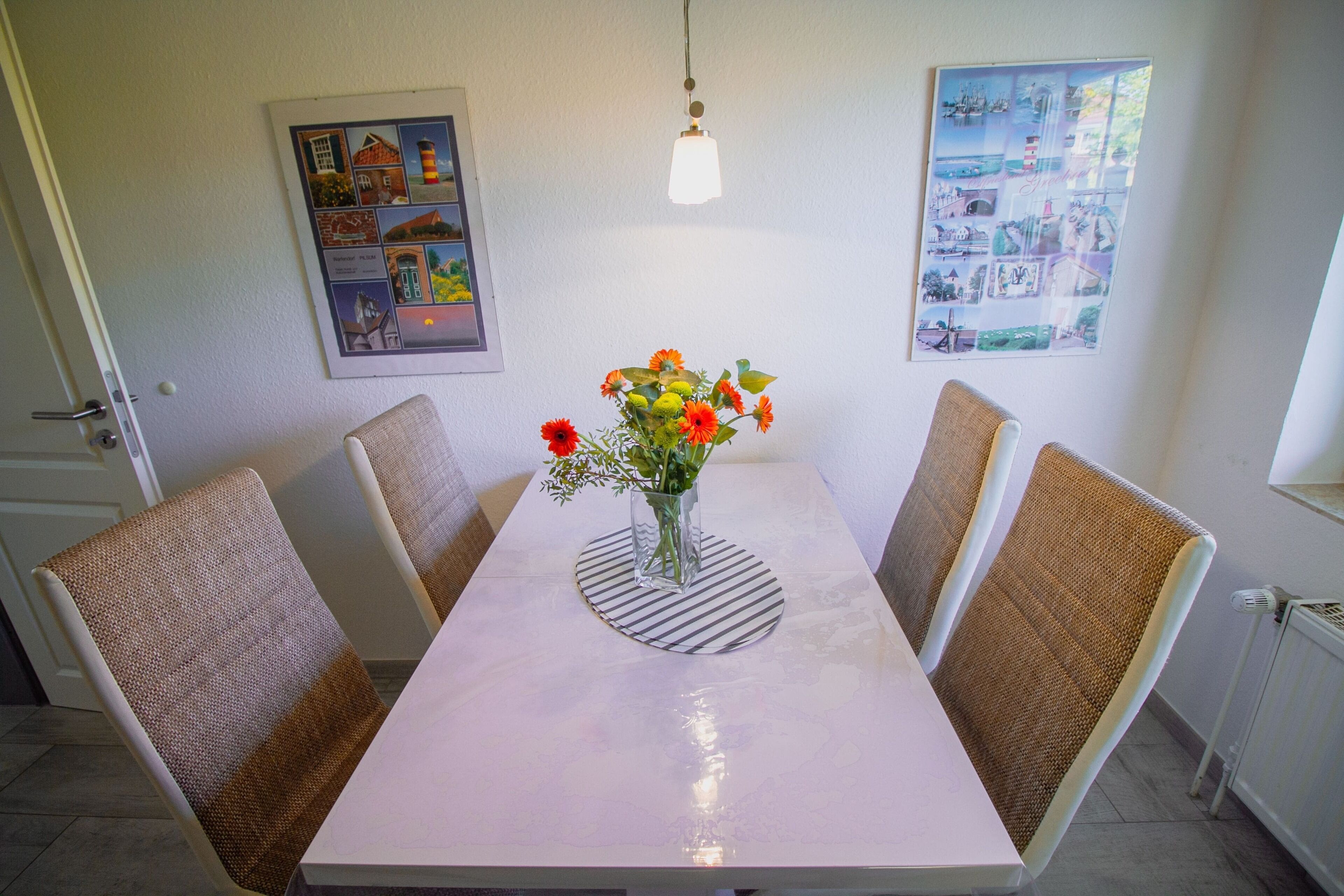 Un comedor con una mesa blanca, cuatro sillas de mimbre, un florero con flores y dos cuadros en la pared.