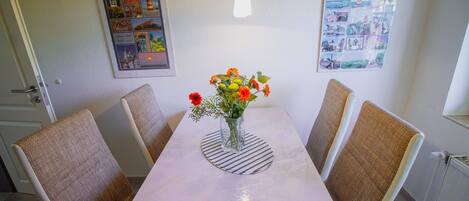 Un comedor con una mesa blanca, cuatro sillas de mimbre, un florero con flores y dos cuadros en la pared.