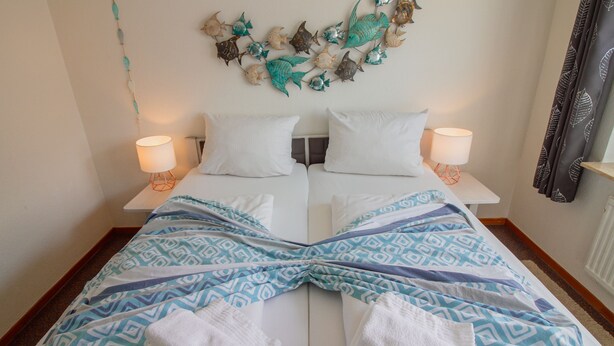 Een tweepersoonsbed met witte lakens en een blauw geborduurd plaid, twee bedlampjes en een wanddecoratie van vissen.