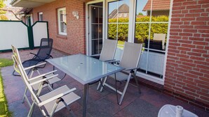 Een terras buiten met een glazen tafel en stoelen, een bakstenen muur en een raam met uitzicht op een huis en begroeiing.