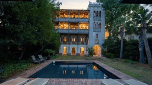 A pool area with lounge chairs, a brick patio, and a building with arched windows and a tower. - Incredible Coastal Castle on Sullivans Island (Sullivans Island)