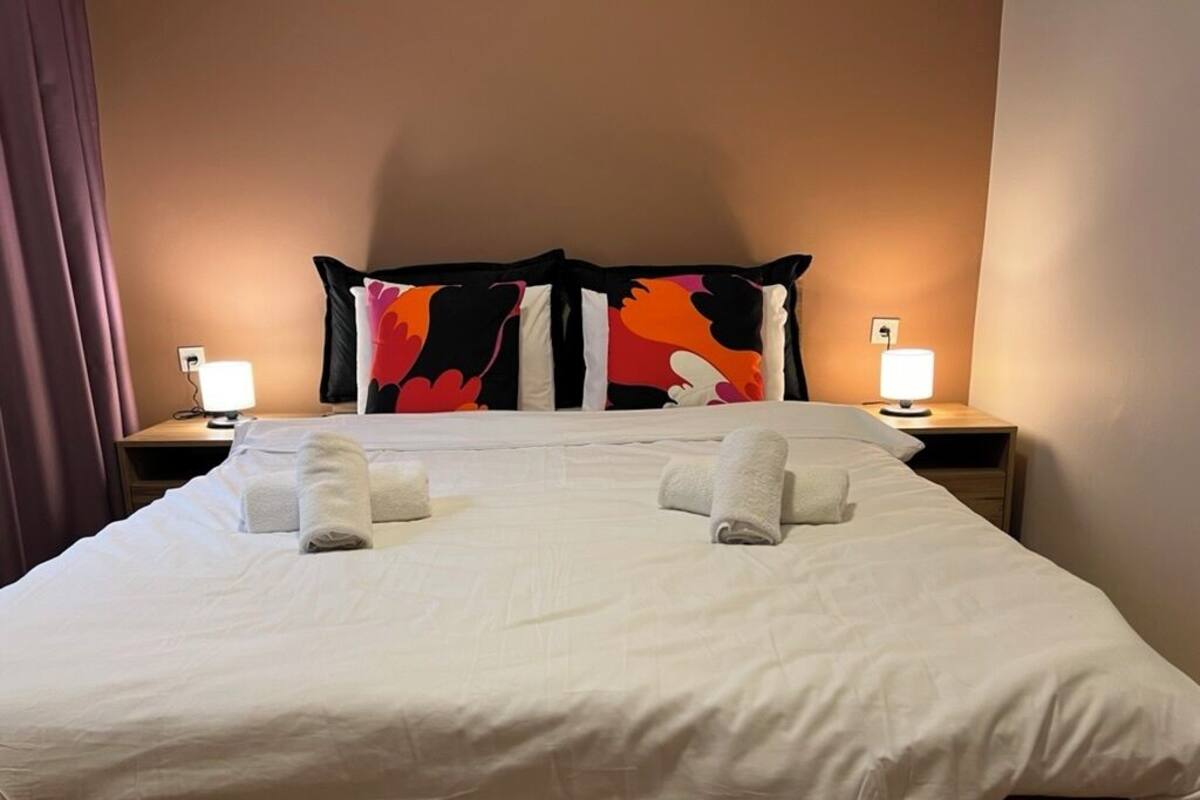 A neatly made bed with white linens and two rolled towels, a colorful abstract pillow, and bedside lamps on wooden nightstands.