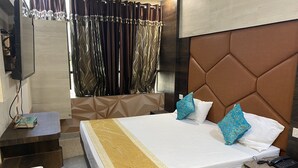 A hotel room with a large bed, a TV mounted on the wall, a bedside table with a phone and remote controls, and a window with curtains. - Hotel Brij Residency (Bhopal)