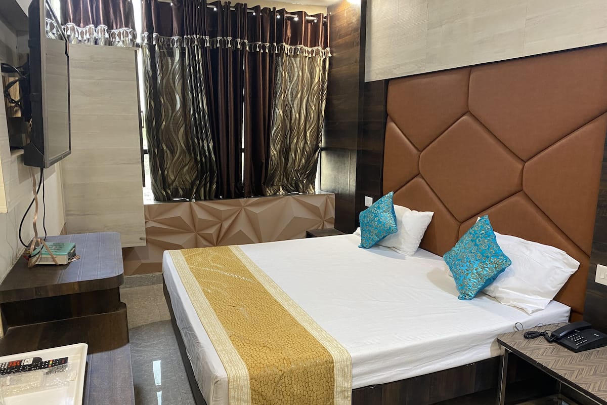 A hotel room with a large bed, a TV mounted on the wall, a bedside table with a phone and remote controls, and a window with curtains.