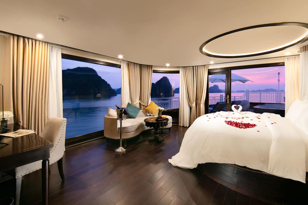 Una lujosa habitación de hotel con una cama grande, una mesa de comedor y vistas a un cuerpo de agua al atardecer.