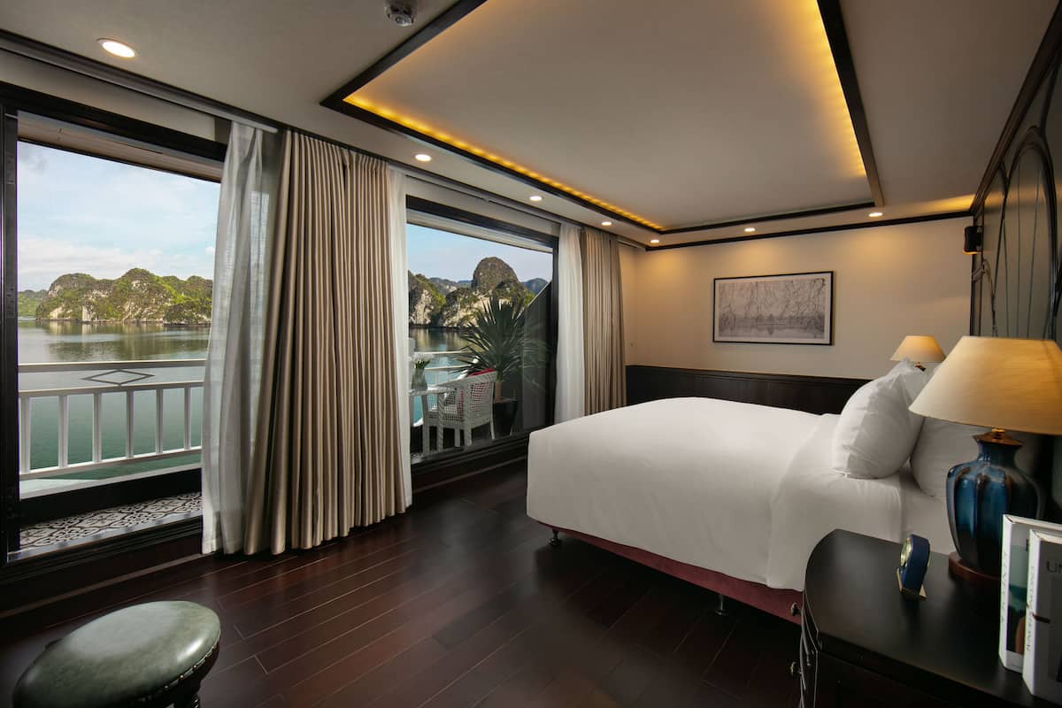 Habitación de hotel con una cama grande, un escritorio, una silla, una ventana con cortinas y vistas a un cuerpo de agua y montañas.