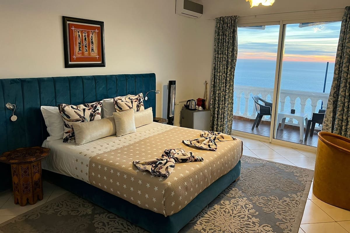 Quarto com uma cama grande, vista para o mar, varanda com cadeiras e um quadro na parede.