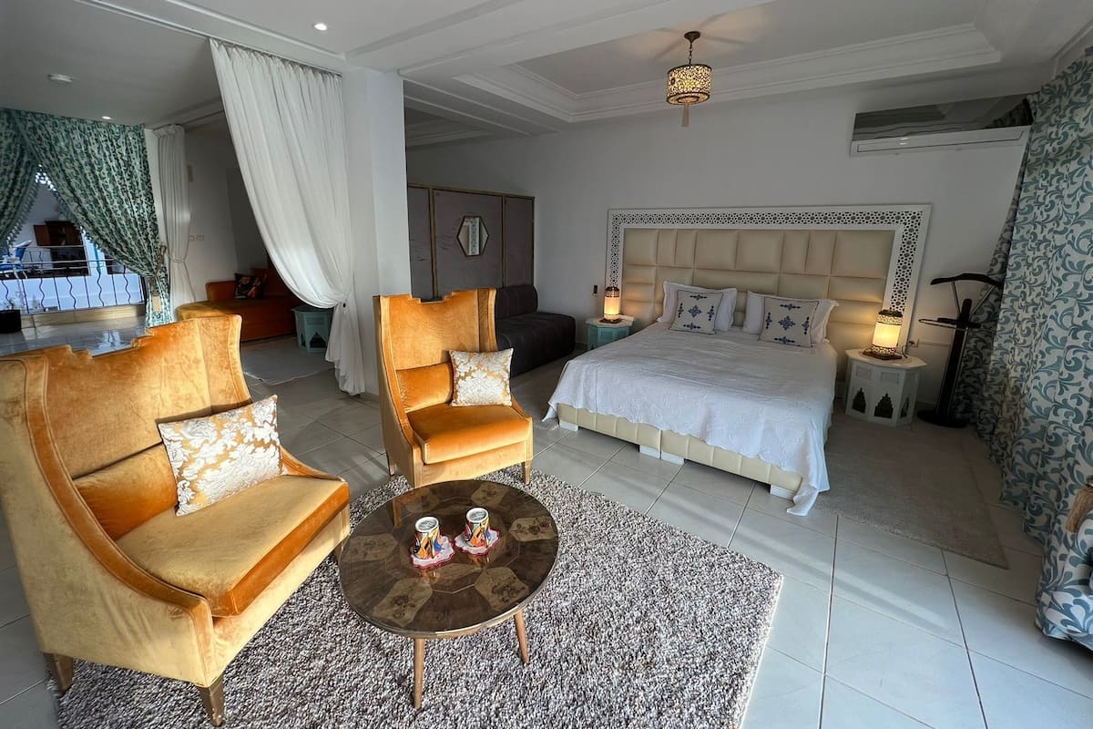 Quarto de hotel com uma cama grande, duas poltronas, uma mesa de centro e vista para o exterior.