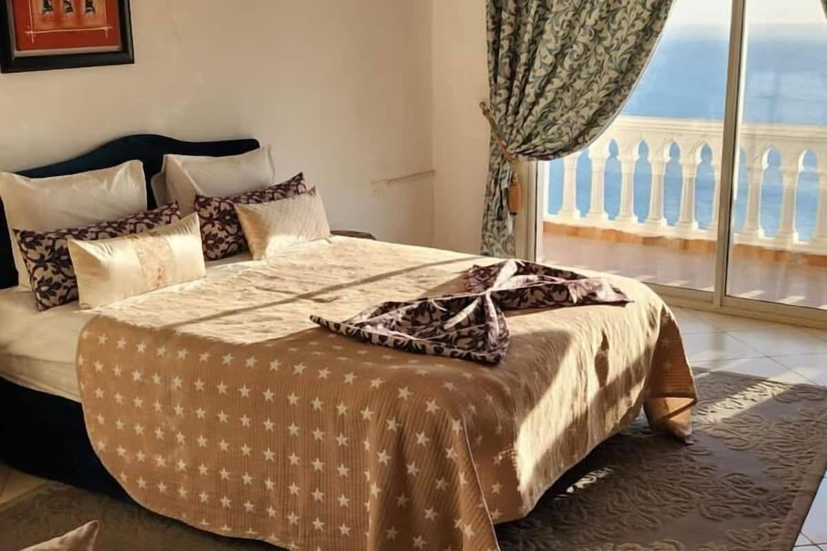 Quarto com cama, almofadas, tapete, janela com cortinas e ar condicionado.
