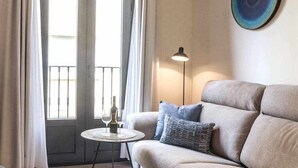 Una sala de estar con un sofá, una mesita redonda con una botella y vasos, una lámpara de pie y vistas a través de una ventana con cortinas.