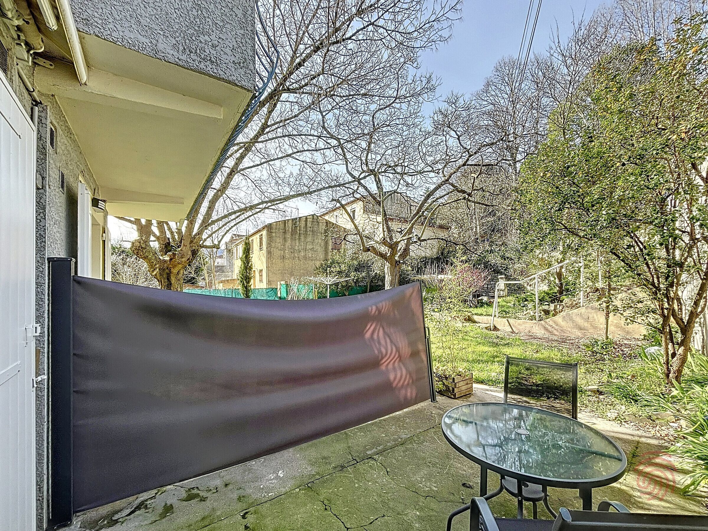 Eine Terrasse mit Glastisch und Stühlen, einer schwarzen Plane und einem Garten mit Bäumen und einem Zaun.