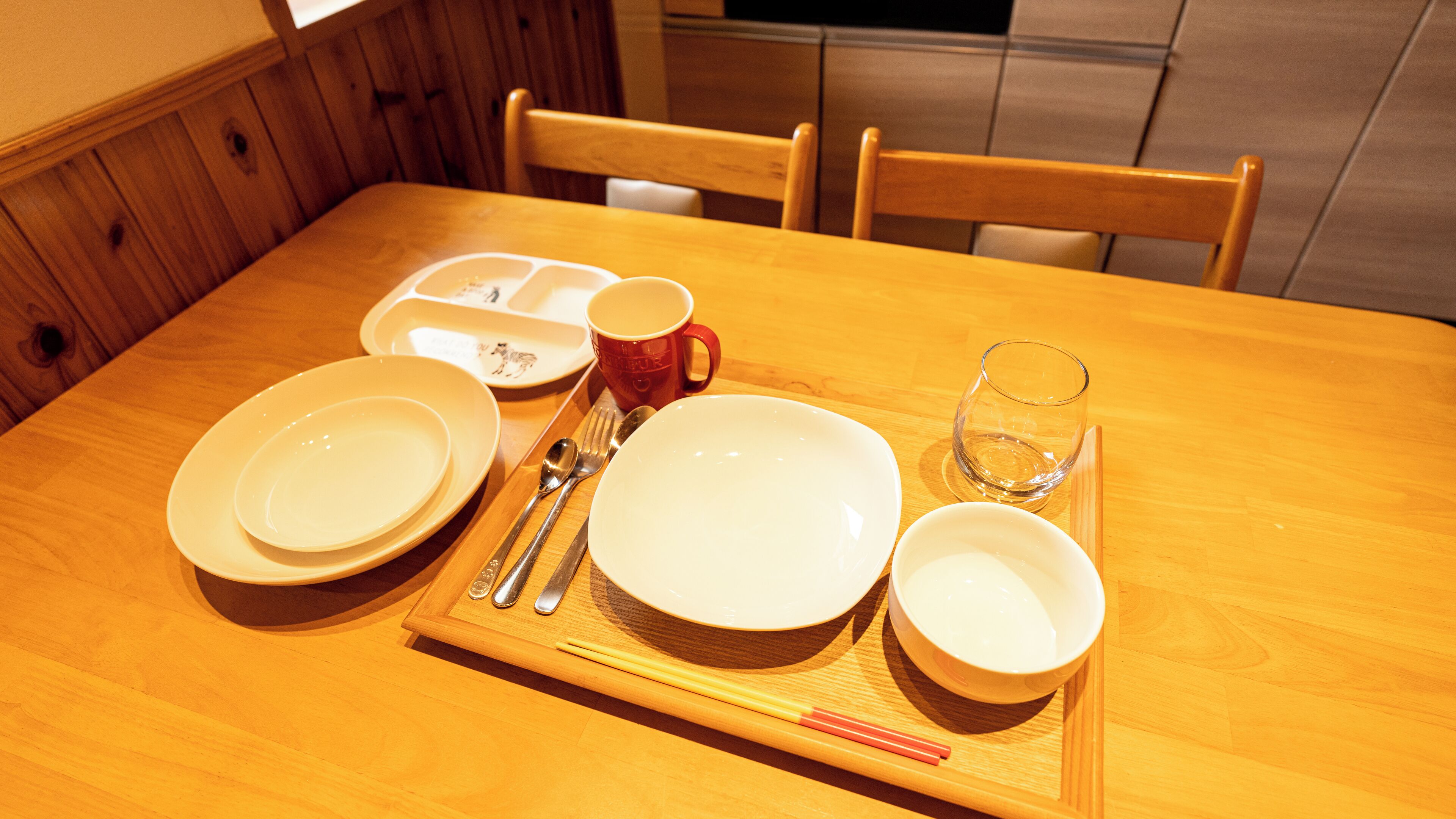 Une table de salle à manger en bois, avec des assiettes, une tasse rouge, des baguettes et un verre.