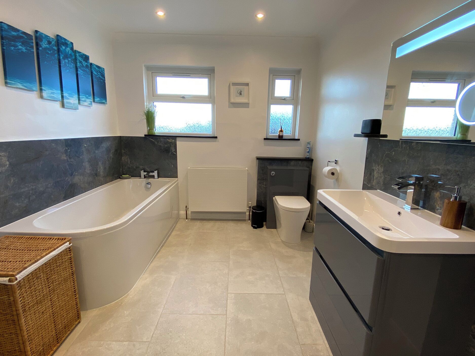 A modern bathroom with a bathtub, a sink, and a toilet. There are two windows providing natural light, and the floor is tiled.