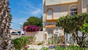 Edifici de dues plantes amb un balcó, un jardí amb diverses plantes i un carrer amb cotxes aparcats.