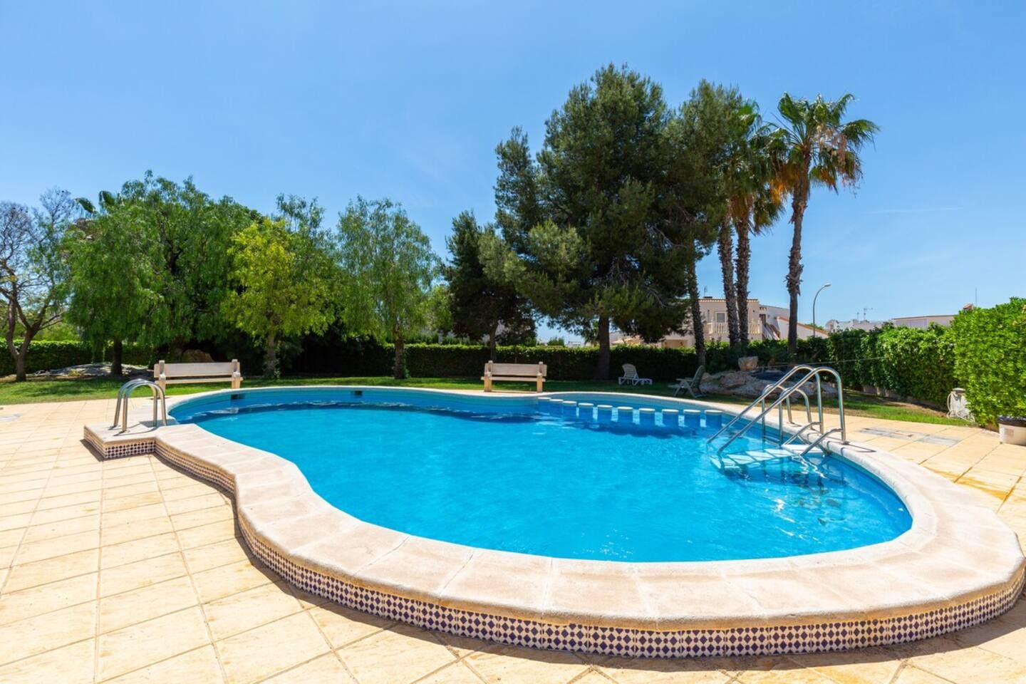 A swimming pool with a tiled deck, surrounded by trees and a clear sky.